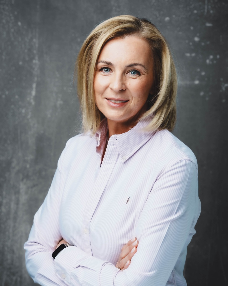 Karolina Specht w jasnej koszuli, z założonymi rękami, na szarym tle